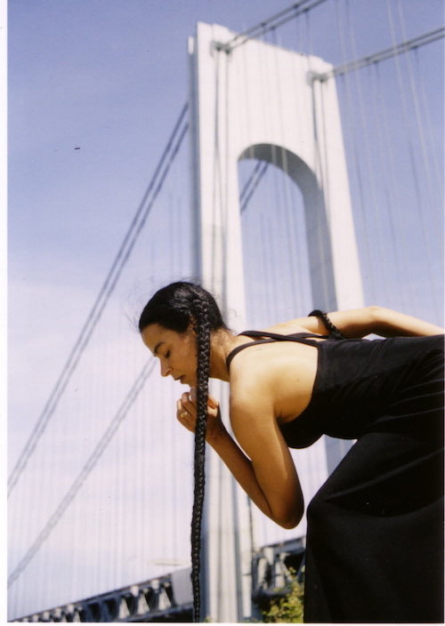 Christa wears a long braid that drapes down to the ground as she assumes a pose. The Brooklyn Bridge is in the background.